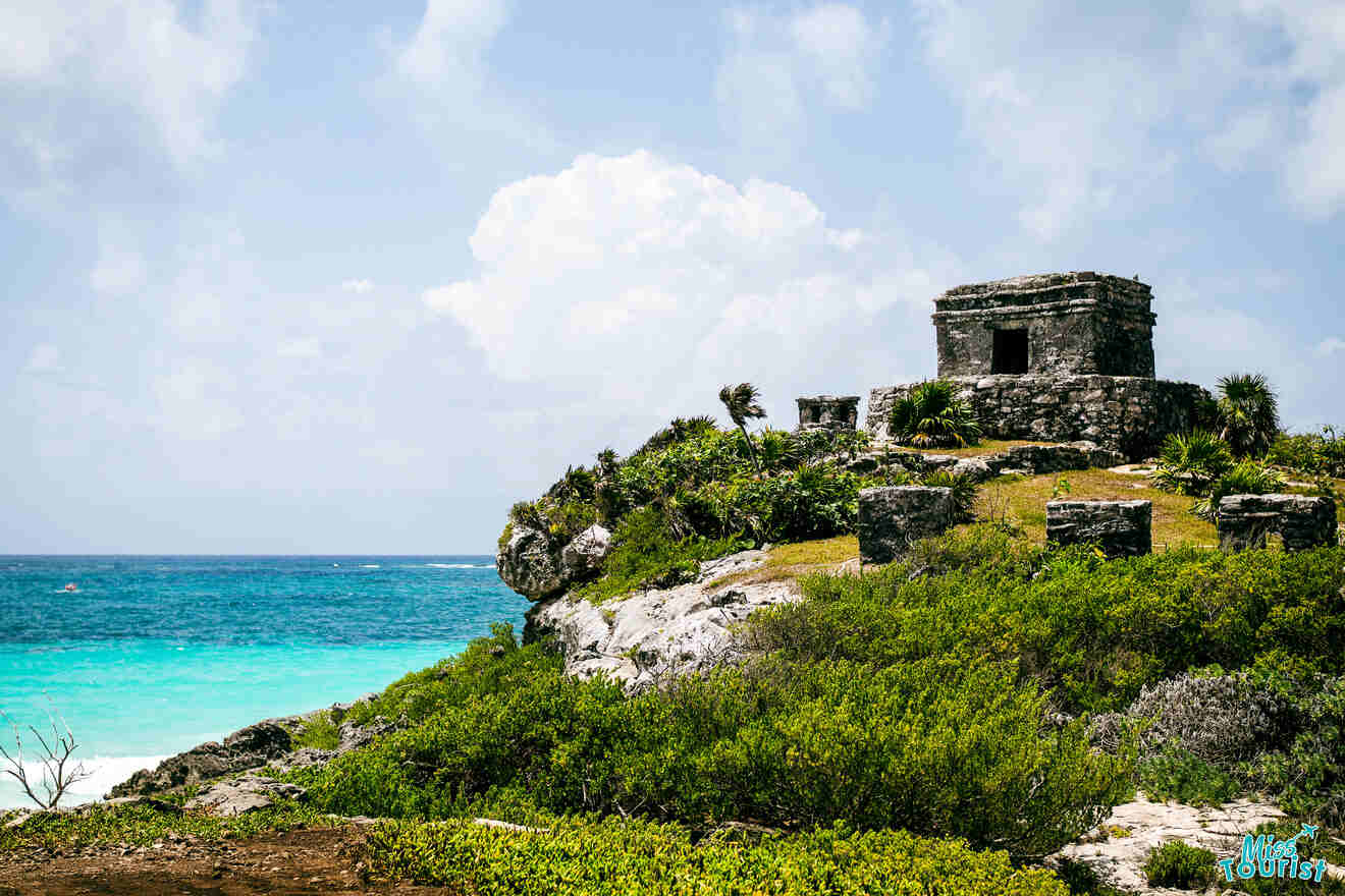 Ancient stone ruins perched on a cliff overlooking a turquoise sea, with a partially cloudy sky above and lush greenery surrounding the structure.