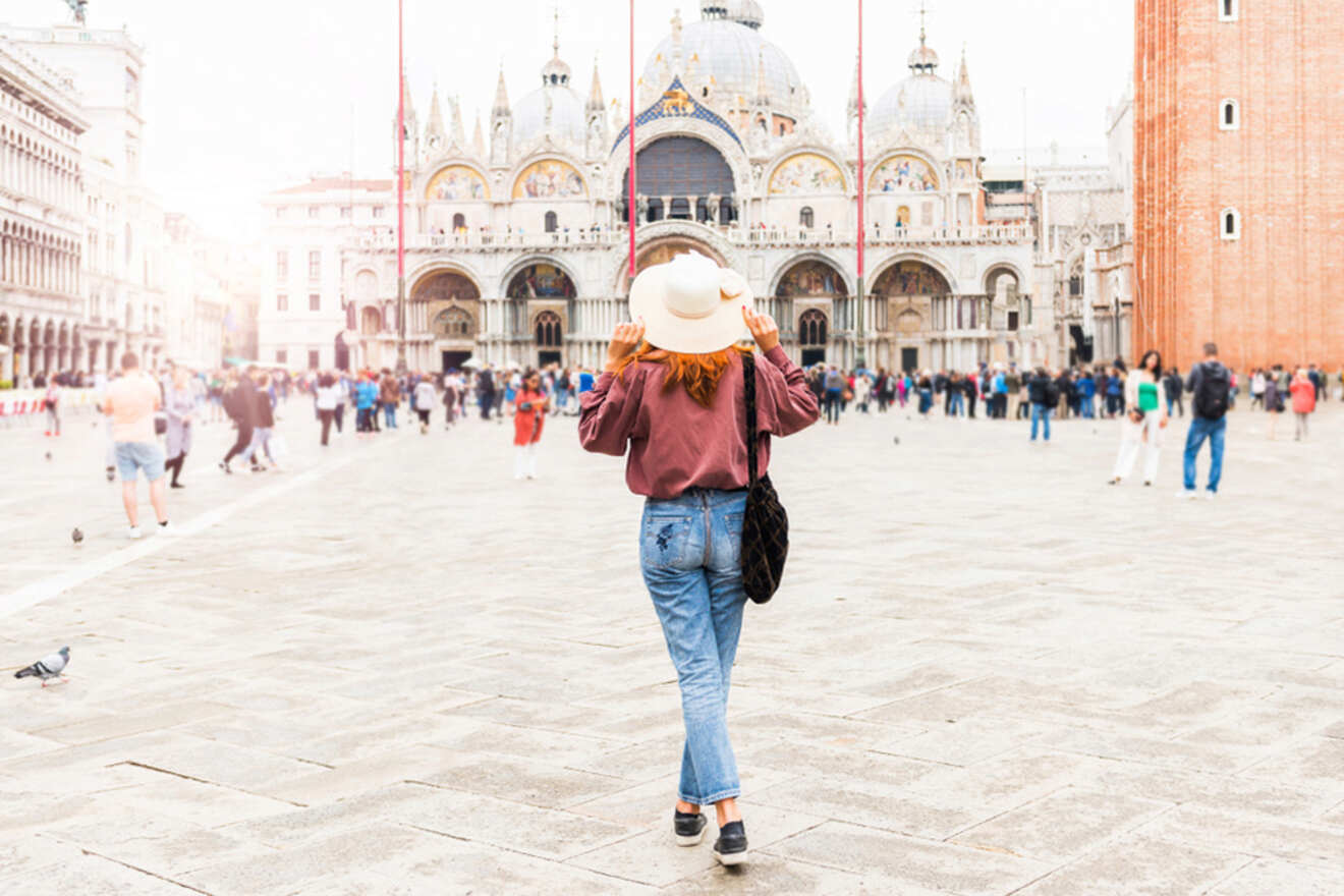 A person wearing a hat and casual clothes walks across a large plaza with a historic building and a tower in the background. The area is populated with other people.