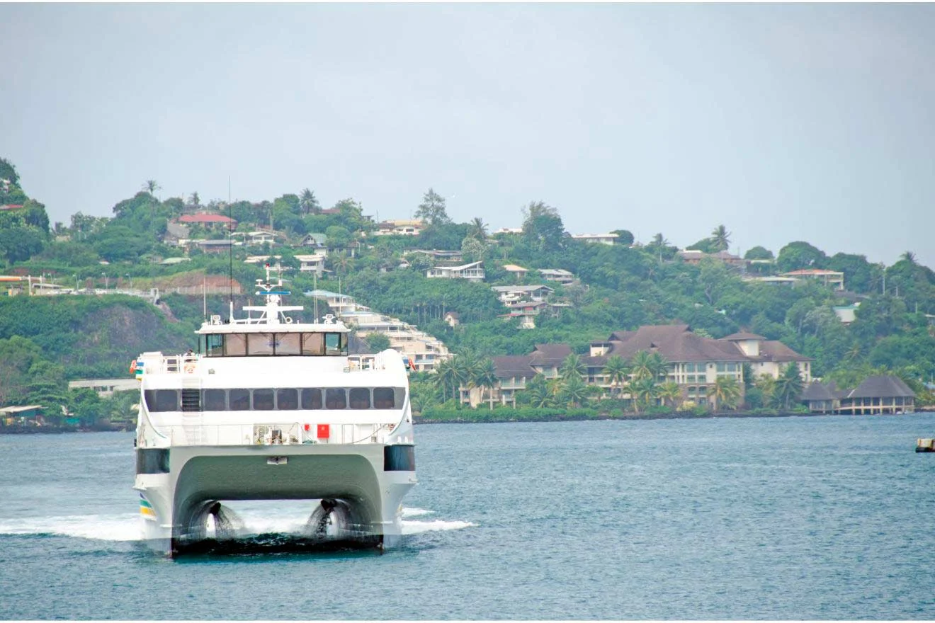 A white catamaran ferry sails across blue water towards the camera with a lush green hilly background.