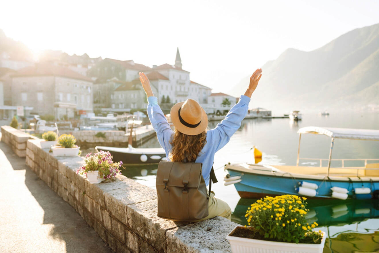 A person with a backpack and hat sits on a stone wall by the water, arms raised. Boats and houses are seen in the background, with mountains in the distance under a bright sky.