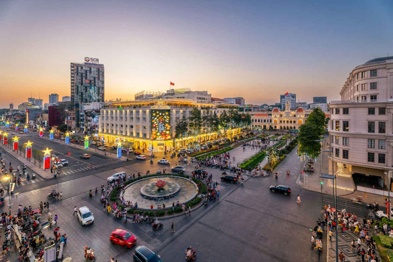 Wide-angle view of a vibrant city square at dusk, featuring a central fountain, busy streets with cars and pedestrians, and a mix of modern buildings and historical architecture.