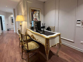 A room with a decorative desk, two ornate chairs, a computer monitor, a lamp, and framed artwork on the wall. The room has wooden flooring and cream-colored walls.