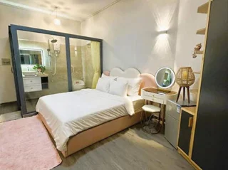 A bedroom with a double bed, vanity table with mirror, and an attached glass-walled bathroom. The room includes shelves, a lamp, and a pink rug.