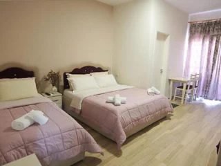 A room with two neatly made double beds with pink bedding. Towels are placed on each bed. A small table and two chairs are by the window with curtain. A door is visible in the background.