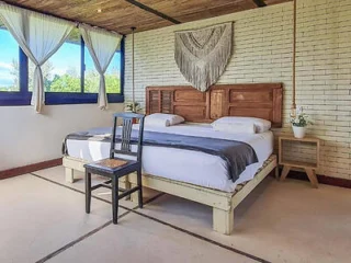 A rustic bedroom with a wooden bed, white bedding, a gray runner, a black chair at the foot of the bed, white brick wall, macramé art, bedside tables with plants, and curtained windows.