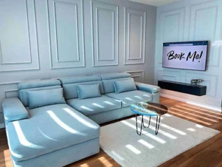 Modern living room with a light blue sectional sofa and a wall-mounted flat-screen TV displaying "Book Me!" A small circular wooden coffee table sits on a white rug, and the walls are paneled.