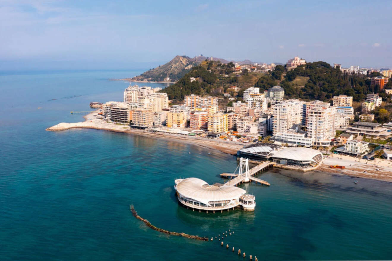 A coastal city with numerous multi-story buildings and hills in the background. A pier extends into the sea, leading to a circular structure surrounded by water.