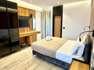 A modern hotel room with a double bed, beige bedding, a wooden desk with a mirror, wall-mounted TV, dark wardrobe, and neutral-colored curtains.