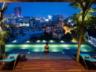 Person sitting at the edge of a rooftop pool at night, overlooking a cityscape with illuminated buildings. Two lounge chairs and trees are positioned on the wooden deck around the pool.