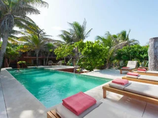 A clear, blue swimming pool surrounded by palm trees and greenery, with four lounge chairs topped with red and white towels placed beside it.