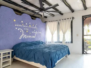 A bedroom with a blue bedspread, white nightstand, purple wall with motivational text, ceiling fan, and white curtains on windows letting in natural light.