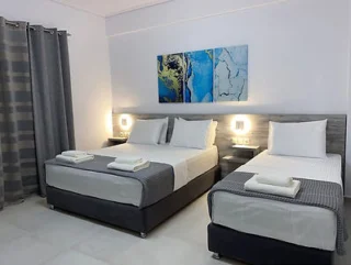 A modern hotel room features two neatly made beds with gray and white bedding, a central nightstand, and abstract blue artwork above the beds. Two wall-mounted lights illuminate the headboard area.