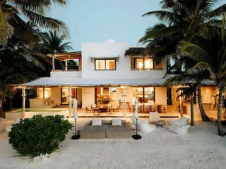 A beachfront house with white walls and palm trees, featuring outdoor seating areas and large windows, set on a sandy beach.