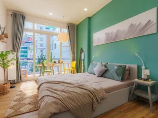 A bright bedroom with a large window, green walls, a bed with gray bedding, a small wooden nightstand, and a dining area with a table and chairs. A modern lamp and decor complete the cozy space.
