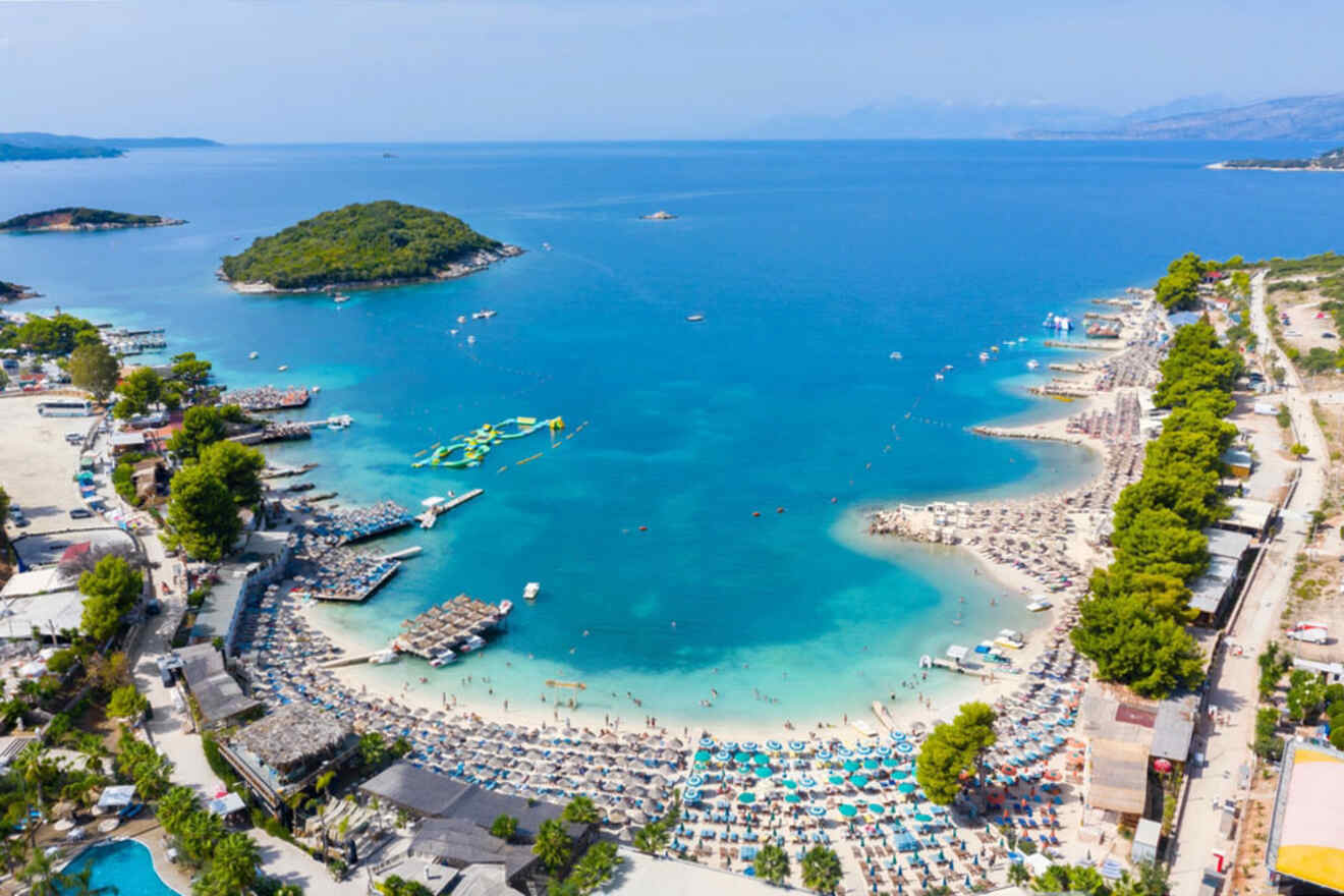 Aerial view of a crowded beach with numerous sun loungers and umbrellas, clear turquoise water, small islands nearby, and surrounding greenery.