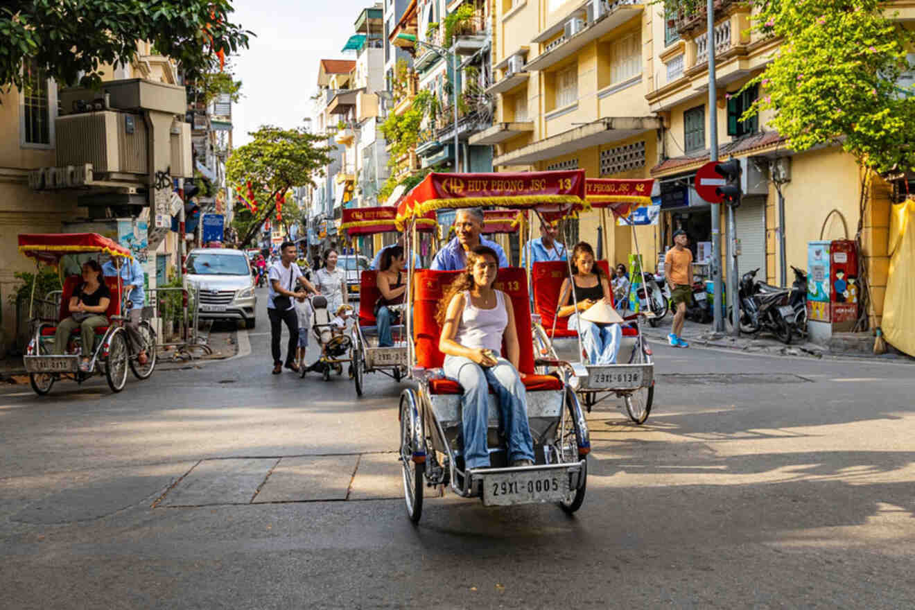People riding traditional rickshaws on a street lined with buildings and trees in a bustling city setting.