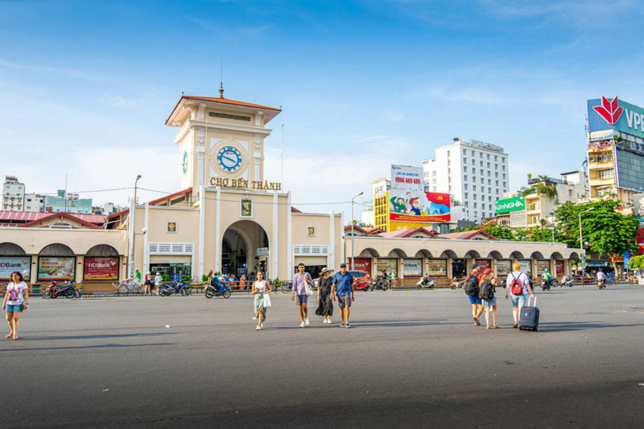People walk and pull luggage across a large street in front of Ben Thanh Market in Ho Chi Minh City, Vietnam, on a sunny day.
