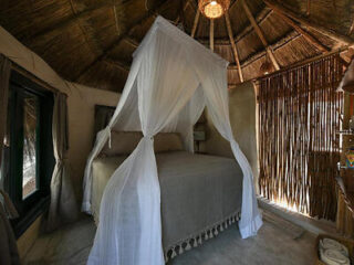 A rustic bedroom with a thatched roof, a canopy bed draped with white netting, and natural light filtering through bamboo blinds.
