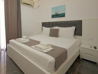 A neatly made bed with a grey headboard and white and grey bedding in a modern room. Two neatly folded towels are placed at the foot of the bed. A painting of a sailboat hangs on the wall above.