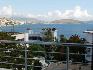 A scenic view from a balcony showing buildings, trees, and a calm body of water with hills in the background under a partly cloudy sky.