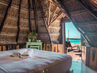 A rustic hut with a thatched roof features a bed with folded towels, a small table with potted plants, and an open door leading to a deck with lounge chairs and an ocean view.