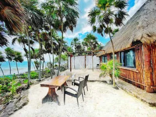 A wooden table and chairs sit on sandy ground near a bamboo hut surrounded by palm trees, with the ocean visible in the background.