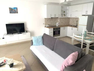 A modern, open-plan living area featuring a grey sofa with pastel cushions, a flat-screen TV, a white kitchen with a dining table, and light-colored tiled flooring.