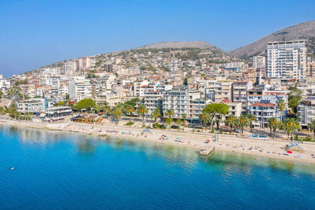 A coastal city with numerous buildings, palm trees along the waterfront, and people on a sandy beach. The clear blue sea is in the foreground, and hills are visible in the background.