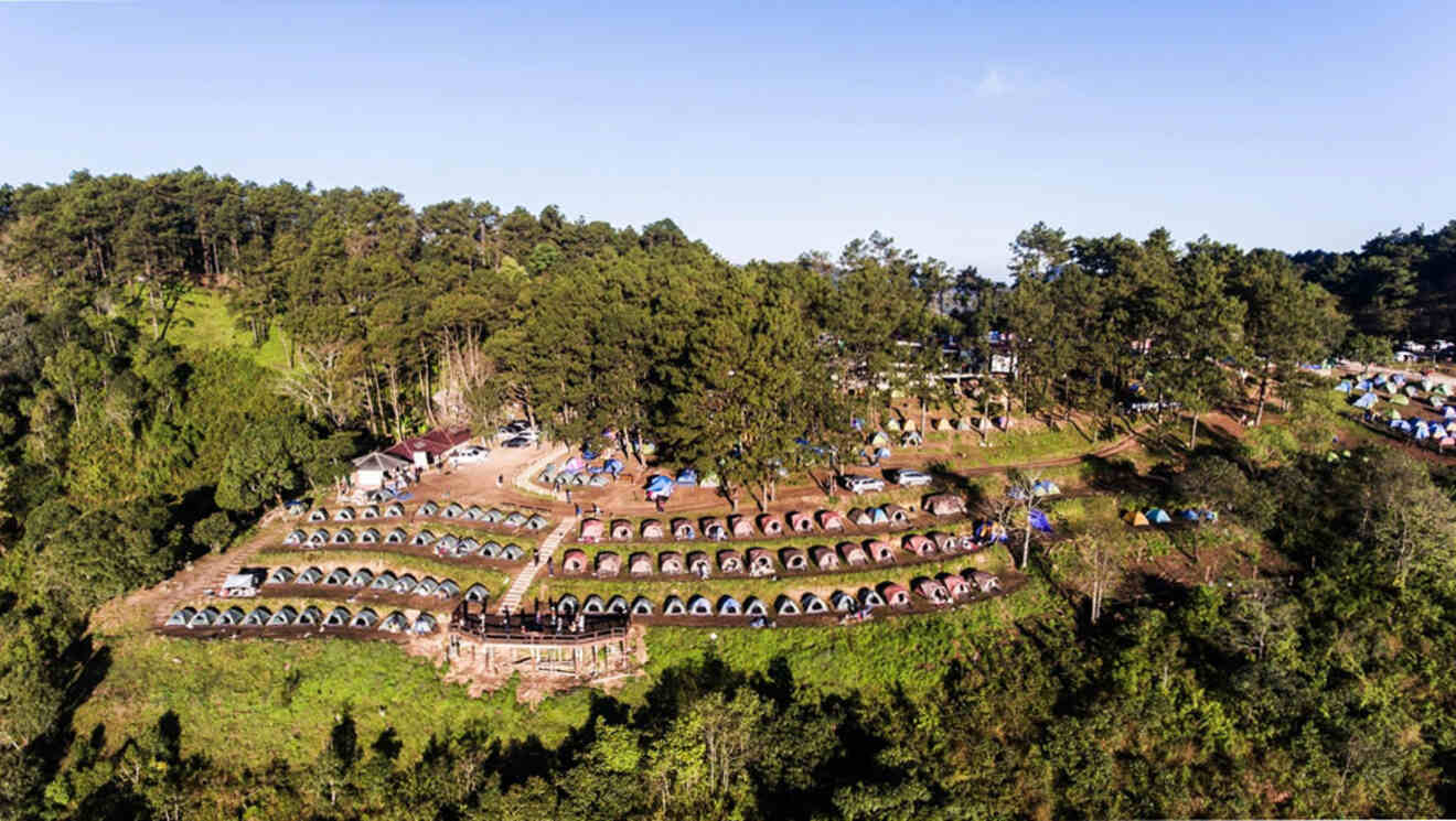 Aerial view of a hillside campground with numerous tents arranged in rows surrounded by dense forest under a clear blue sky.