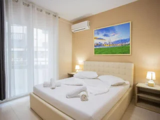 A modern bedroom with a large bed, beige walls, and white curtains. The room features bedside tables with lamps, an air conditioner above the bed, and a framed landscape painting on the wall.