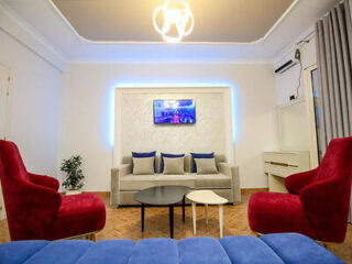 Bright living room with modern furniture. Gray sofa with blue cushions, two red chairs, blue ottoman, black and white coffee tables, wall-mounted TV, indoor plant, and a unique ceiling light fixture.