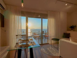 A modern dining area in an apartment, featuring a glass table set for two, with a view of a sunlit balcony and the ocean in the background.