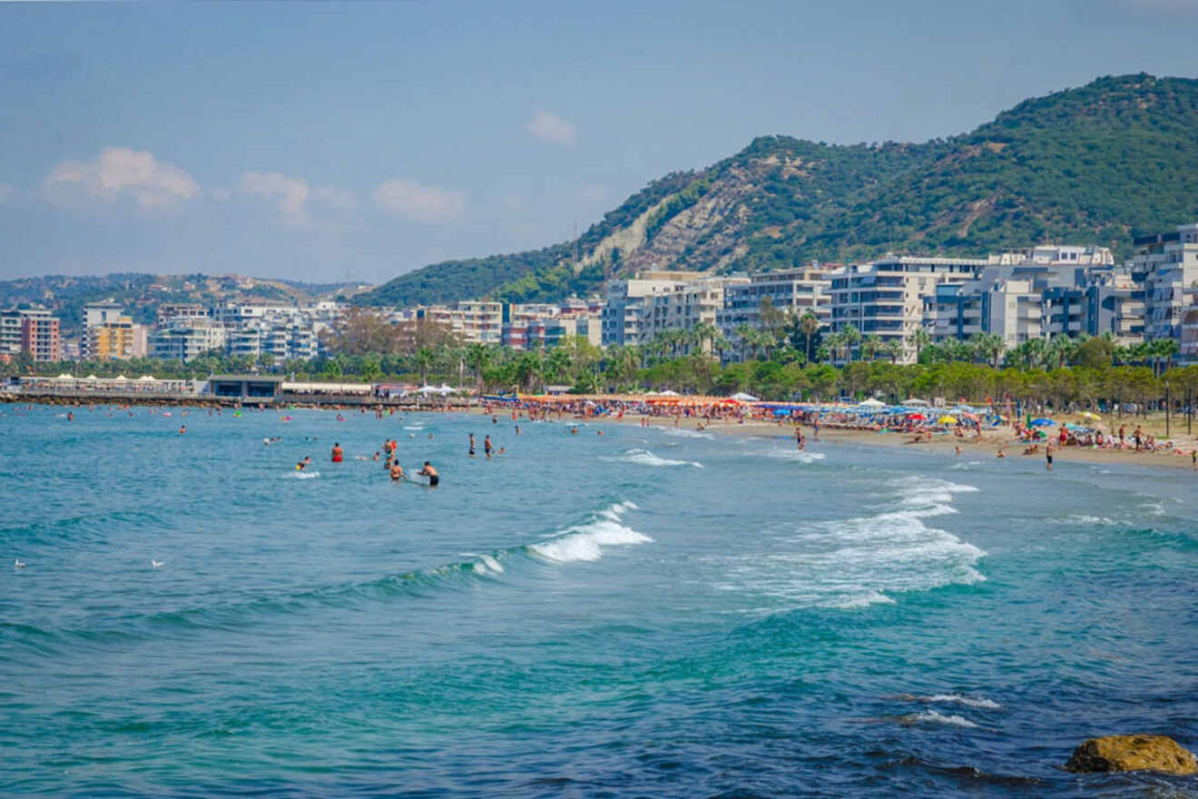 A beach lined with people enjoying the water and sun. Residential buildings and green hills are in the background.