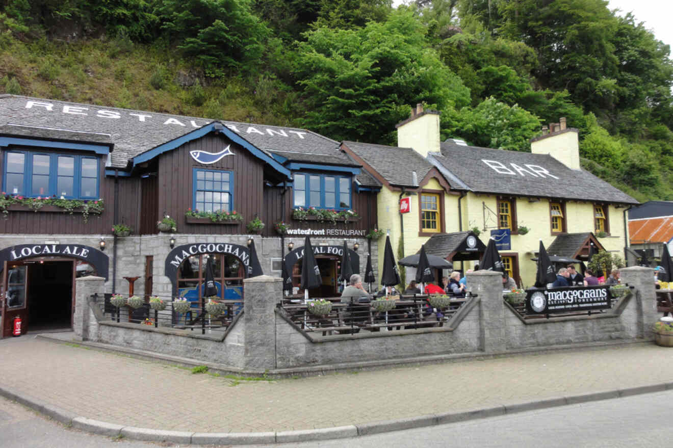 Exterior view of a rustic restaurant and bar with outdoor seating, labeled Macgochans, next to a large sign reading "LOCAL ALES." The restaurant is surrounded by woods and greenery.