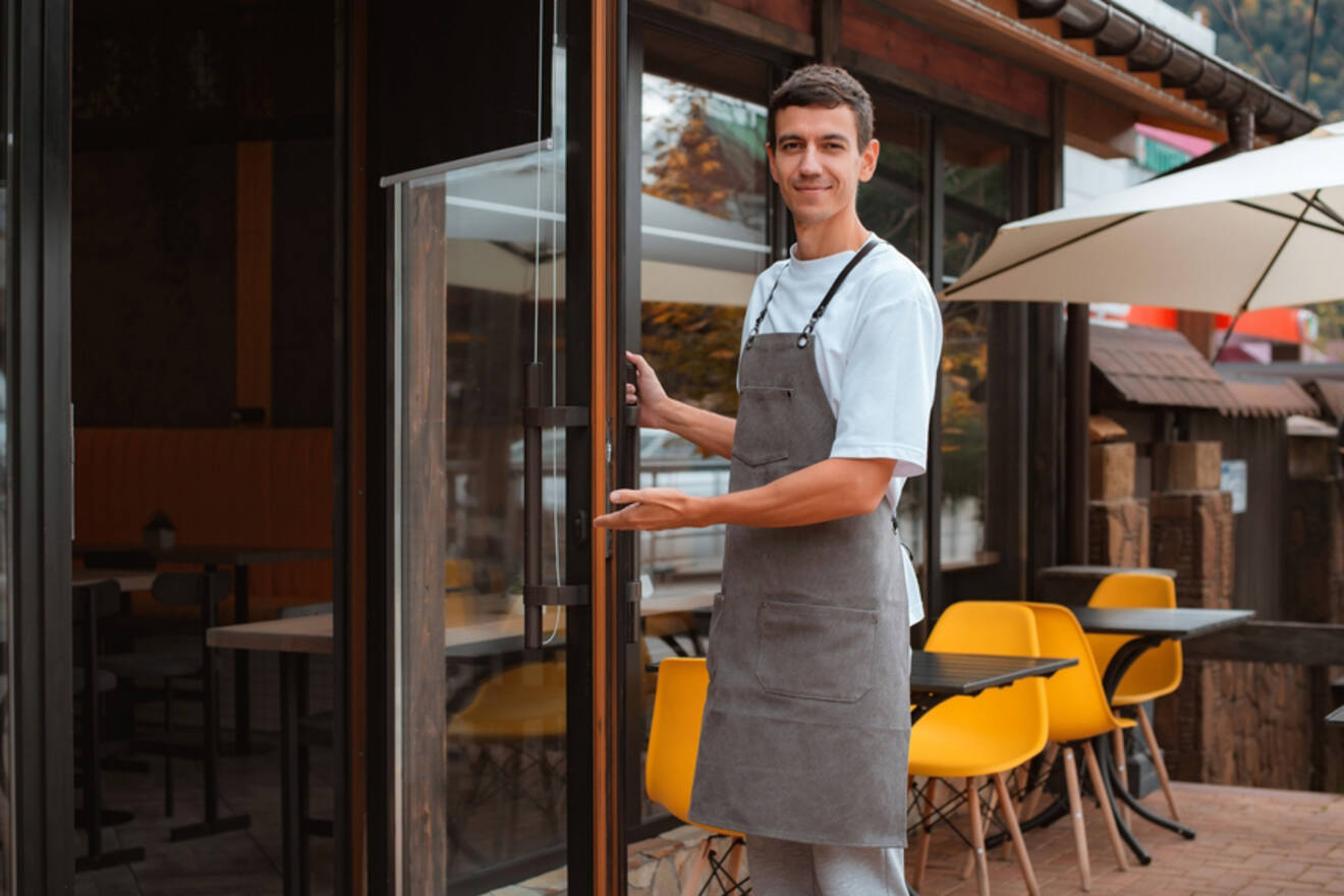 A smiling waiter in an apron stands at the entrance of a restaurant, holding the door open and gesturing for guests to enter.