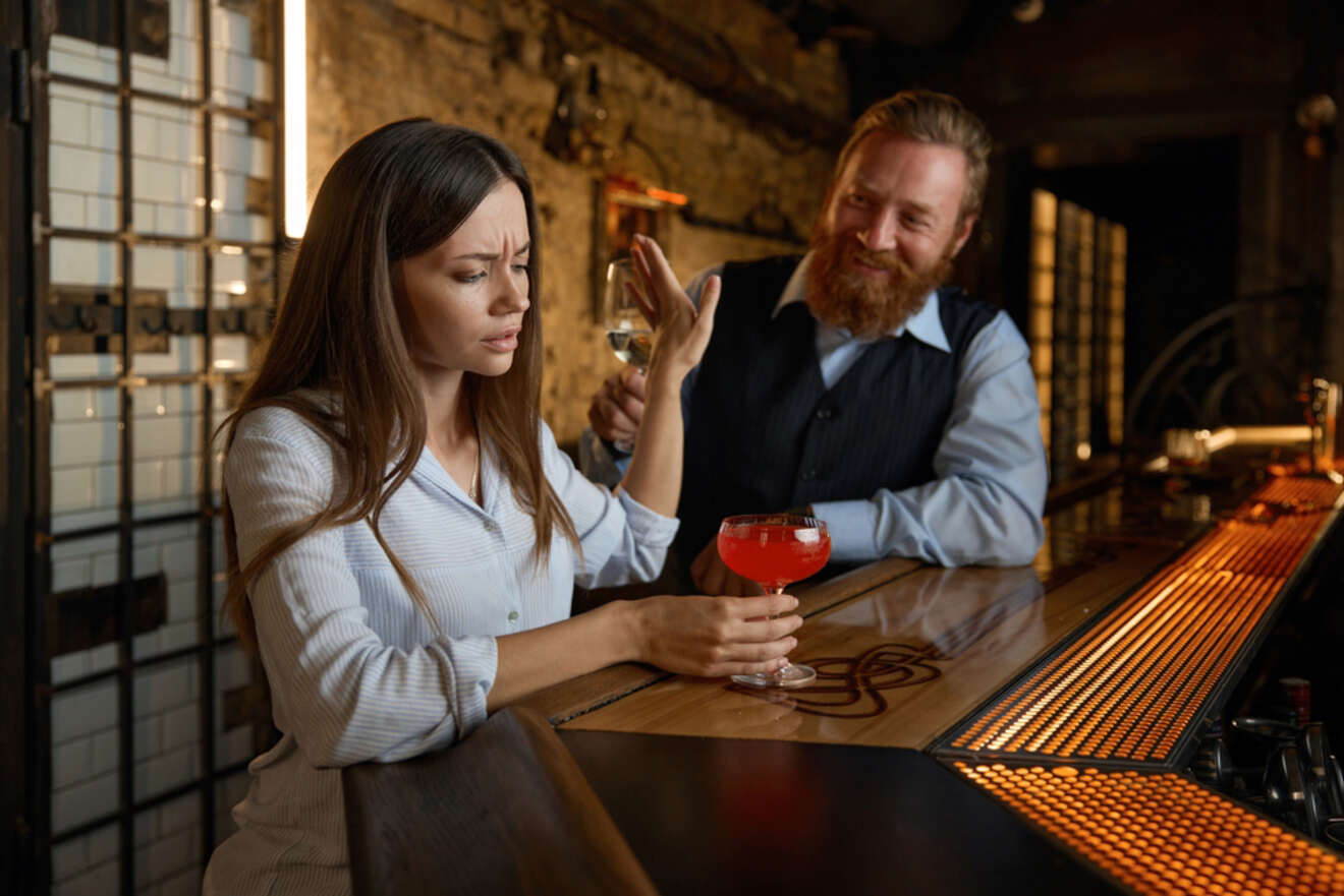 A woman with a displeased expression waves off a drink offered by a bearded man at a bar, the woman holding a red cocktail.