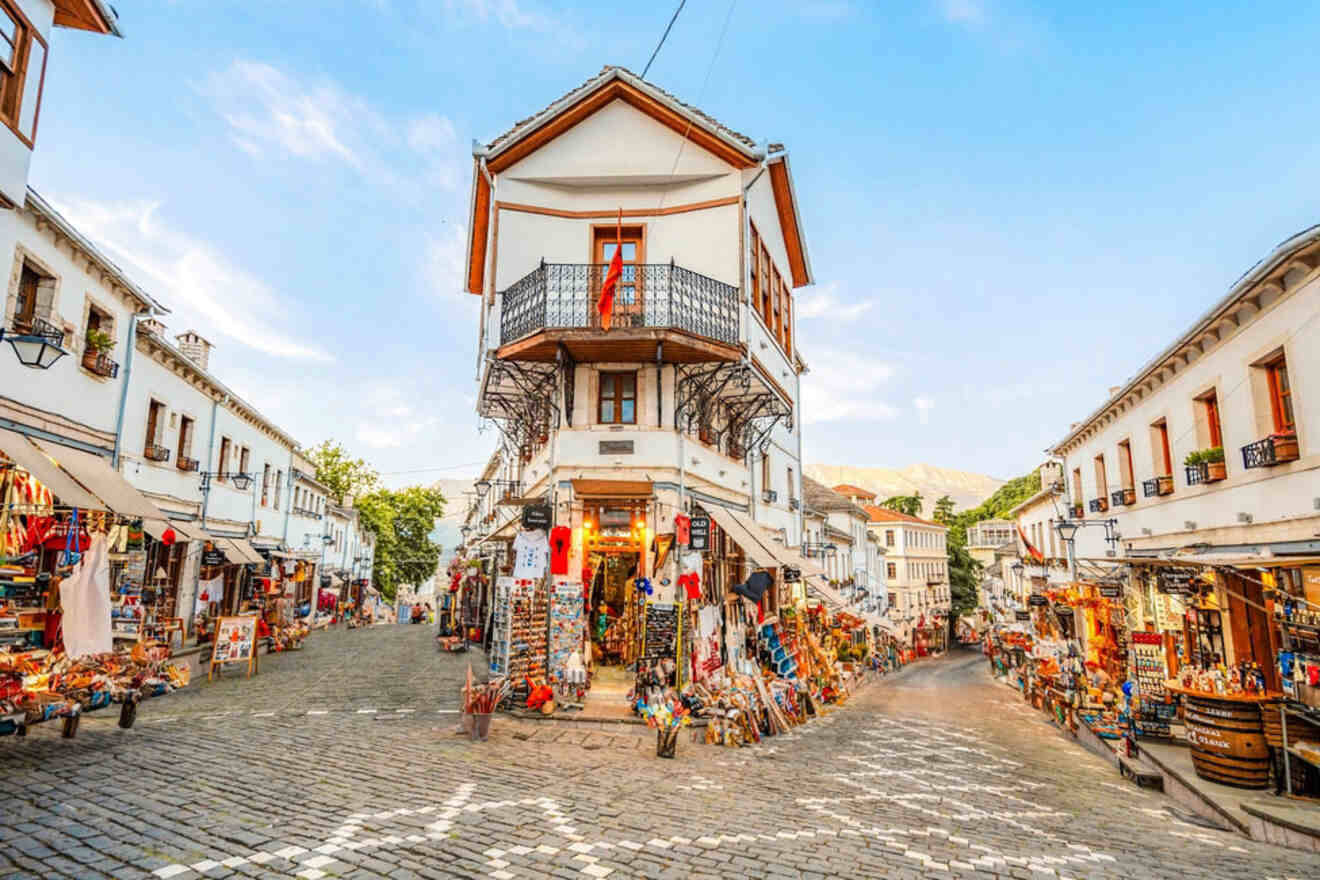 A bustling street in Gjirokaster, lined with white-washed buildings, shops, and a distinctive house with a balcony at the center.