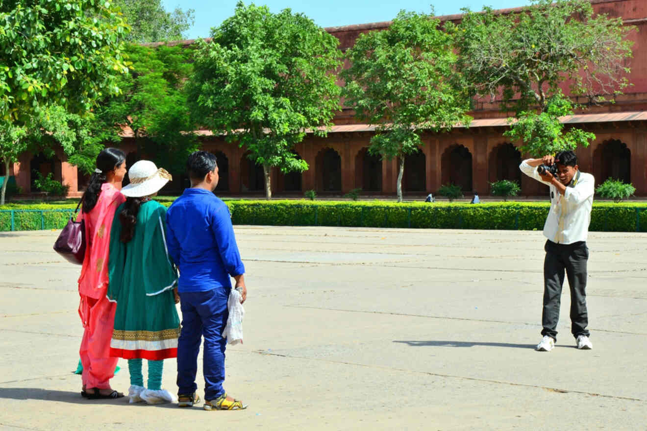 A photographer taking a picture of a group of tourists in traditional Indian attire in front of a historic building, with green trees in the background.