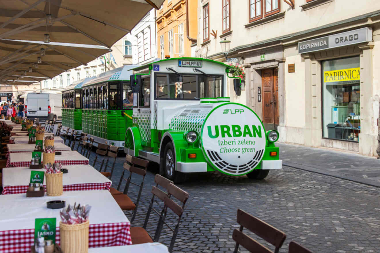 A green and white tourist train labeled "URBAN" travels down a cobblestone street lined with outdoor seating at cafes. The surroundings feature historical buildings.