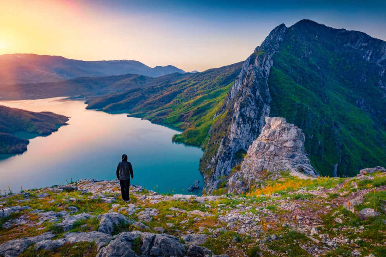 A person stands on a rocky outcrop overlooking the serene Lake Bovilla at sunset, with mountains in the background.