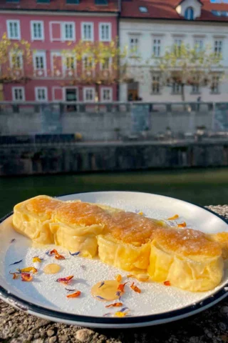 Four rolls of sweet pastry on a white plate with a decorative sprinkle, placed outdoors with colorful buildings and a canal in the background.