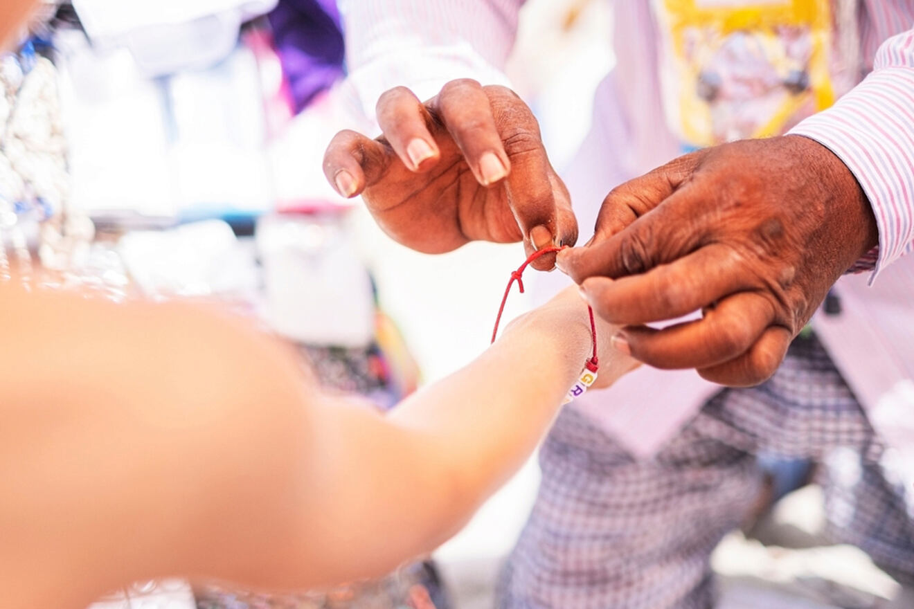 A close-up of a vendor tying a colorful bracelet onto a customer's wrist, the vendor's hands visible against a blurred background of market items.