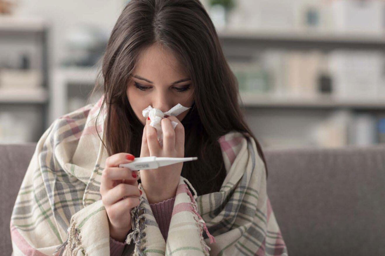 A woman sitting on a couch, wrapped in a blanket, blowing her nose and checking a thermometer.