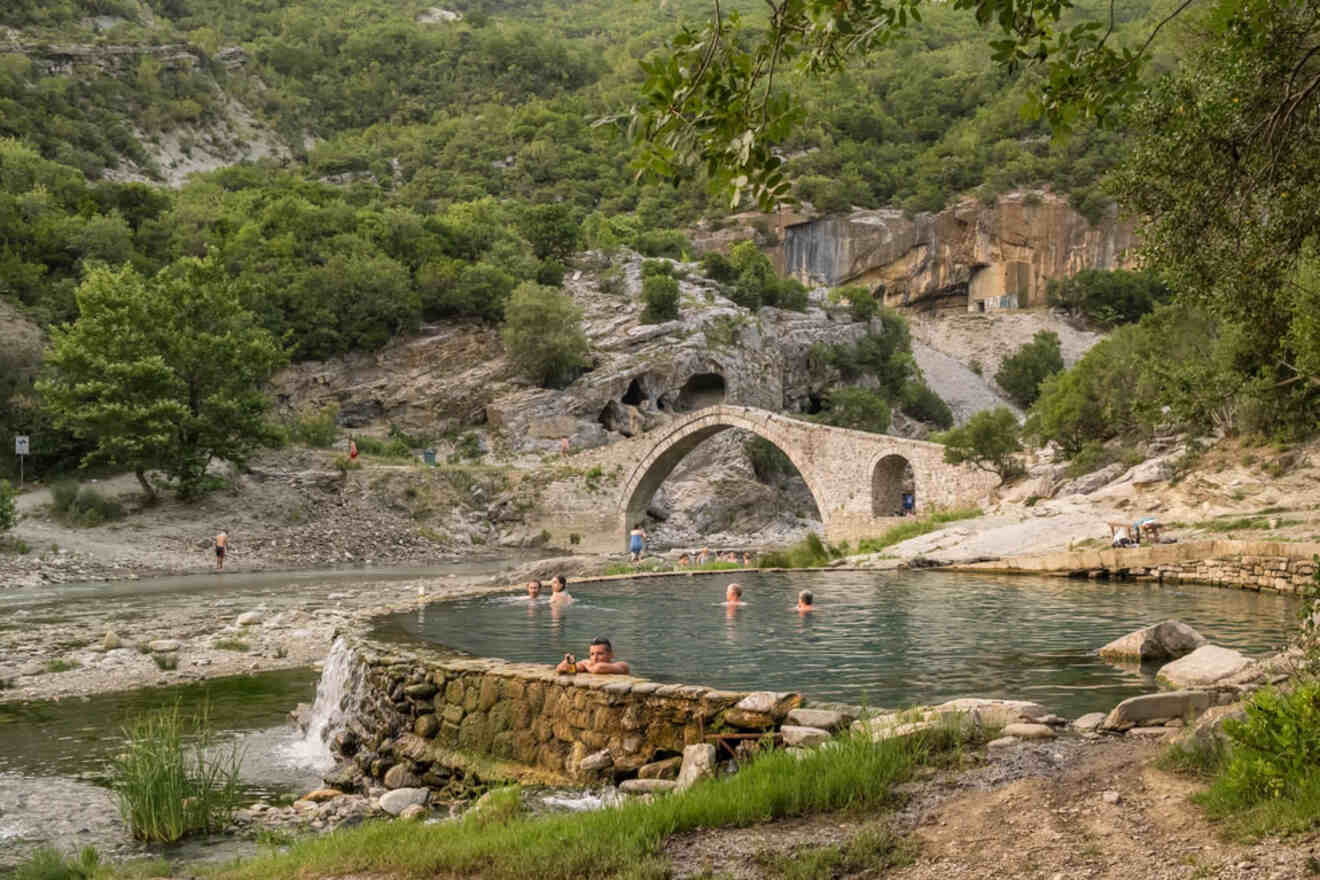 People relax in the natural thermal springs of Permet, with a historic stone bridge and lush landscape in the background.