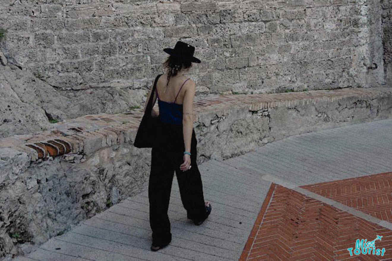 The writer of the post dressed in black with a hat walks along a pathway next to a historic stone wall in Monaco.