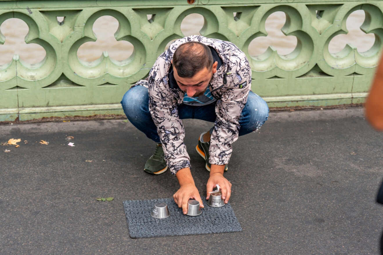 A street performer crouching on the ground, arranging cups for a ball-and-cup game, with a decorative bridge railing in the background.