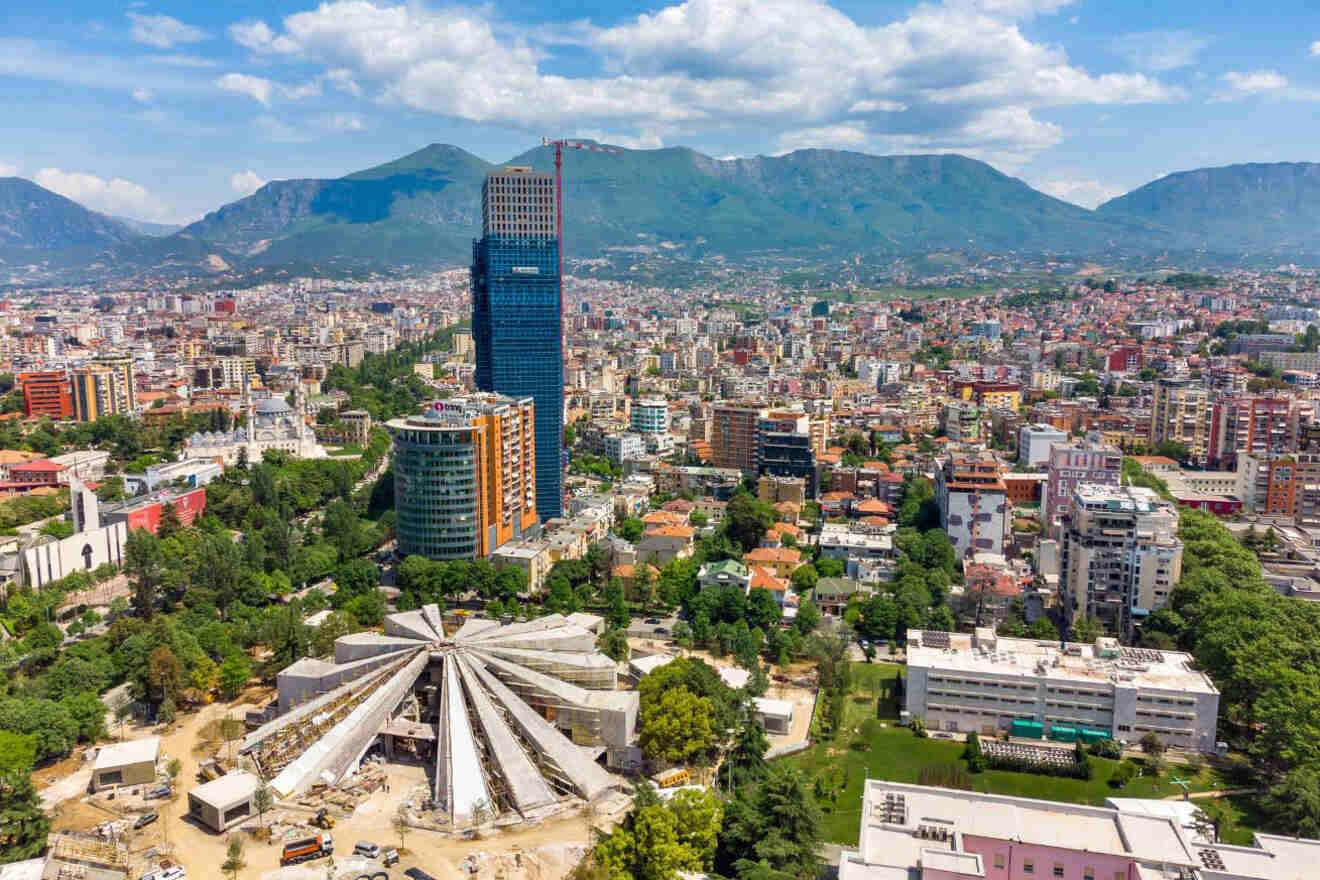 An aerial view of Tirana, featuring modern skyscrapers, historic buildings, and the mountains in the distance.