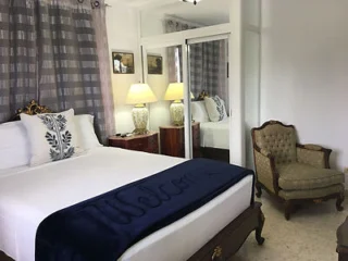 A neatly arranged bedroom with a double bed, decorative pillows, and a dark blue blanket that says "Welcome." There is a cushioned armchair, mirrored closet doors, curtains, and bedside lamps.