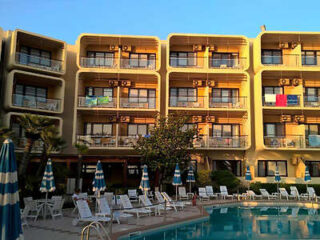 A multi-story hotel with balconies overlooks a swimming pool area surrounded by lounge chairs and striped umbrellas.