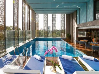 Indoor pool area with blue lounge chairs, folded towels, and a flower pot on a wooden deck. The pool is surrounded by large ornamental windows and has a cityscape view in the background.